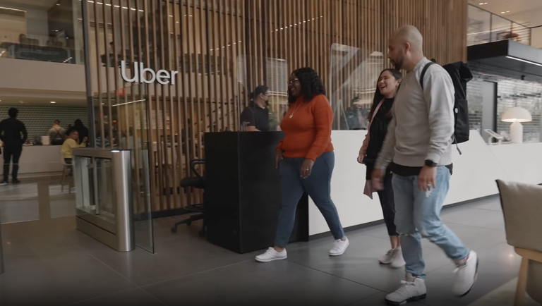 Modern office interior featuring the Uber logo on a wood-slatted partition wall. Three people are visible in business casual attire: a person in an orange sweater walking past a dark reception desk, and two others in light-colored clothing in the background. The space has polished floors, modern white seating, and natural lighting. At the bottom, black text with an arrow icon reads "Uber scales inbound support responses with WRITER".